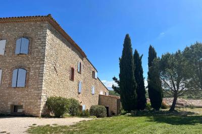Bâtisse en pierre avec toit en tuiles, entourée de végétation et d'un chemin sous un ciel bleu clair.
