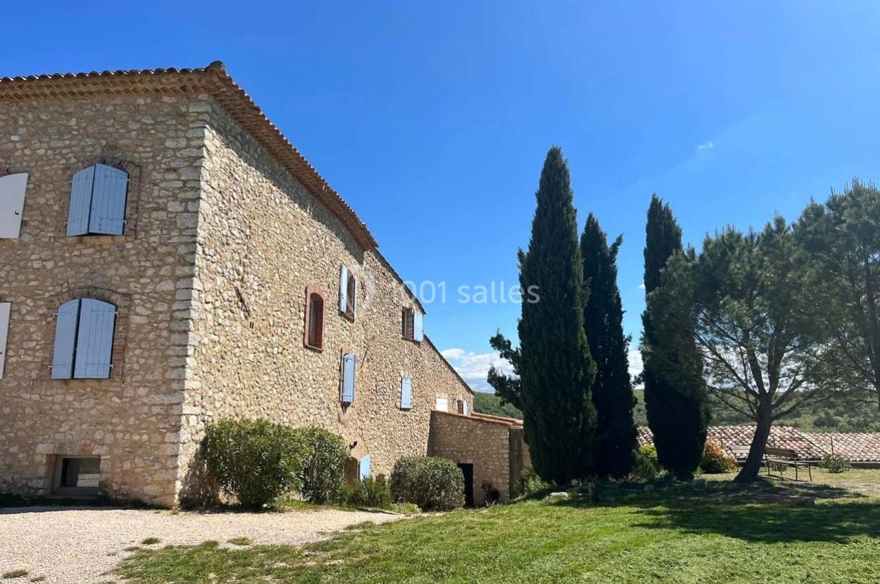 Bâtisse en pierre avec volets blancs, entourée de cyprès et d'arbres, sous un ciel bleu dégagé.