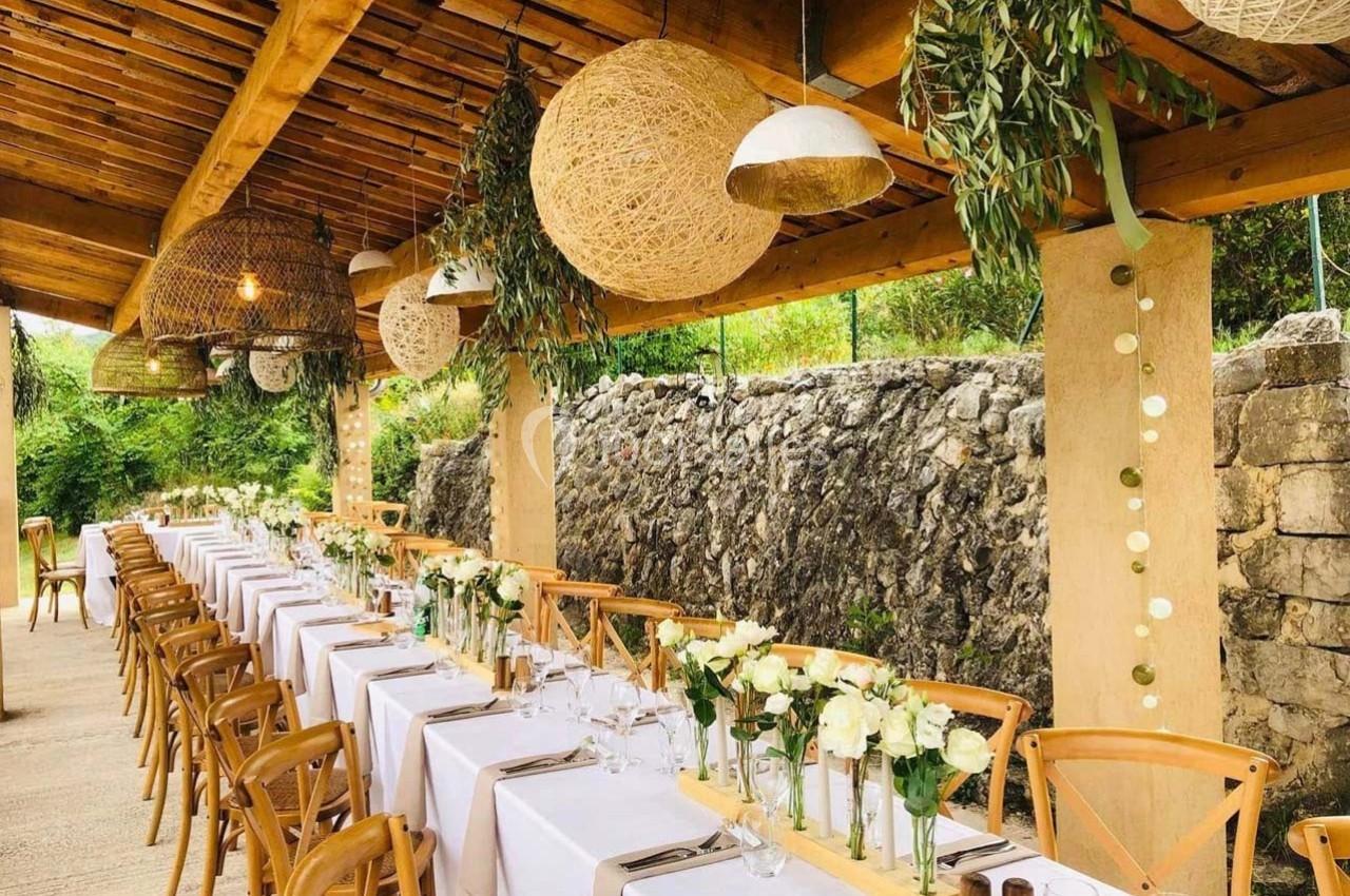 Tables longues décorées de nappes blanches, fleurs et chaises en bois sous une pergola ornée de lampes et feuillages.