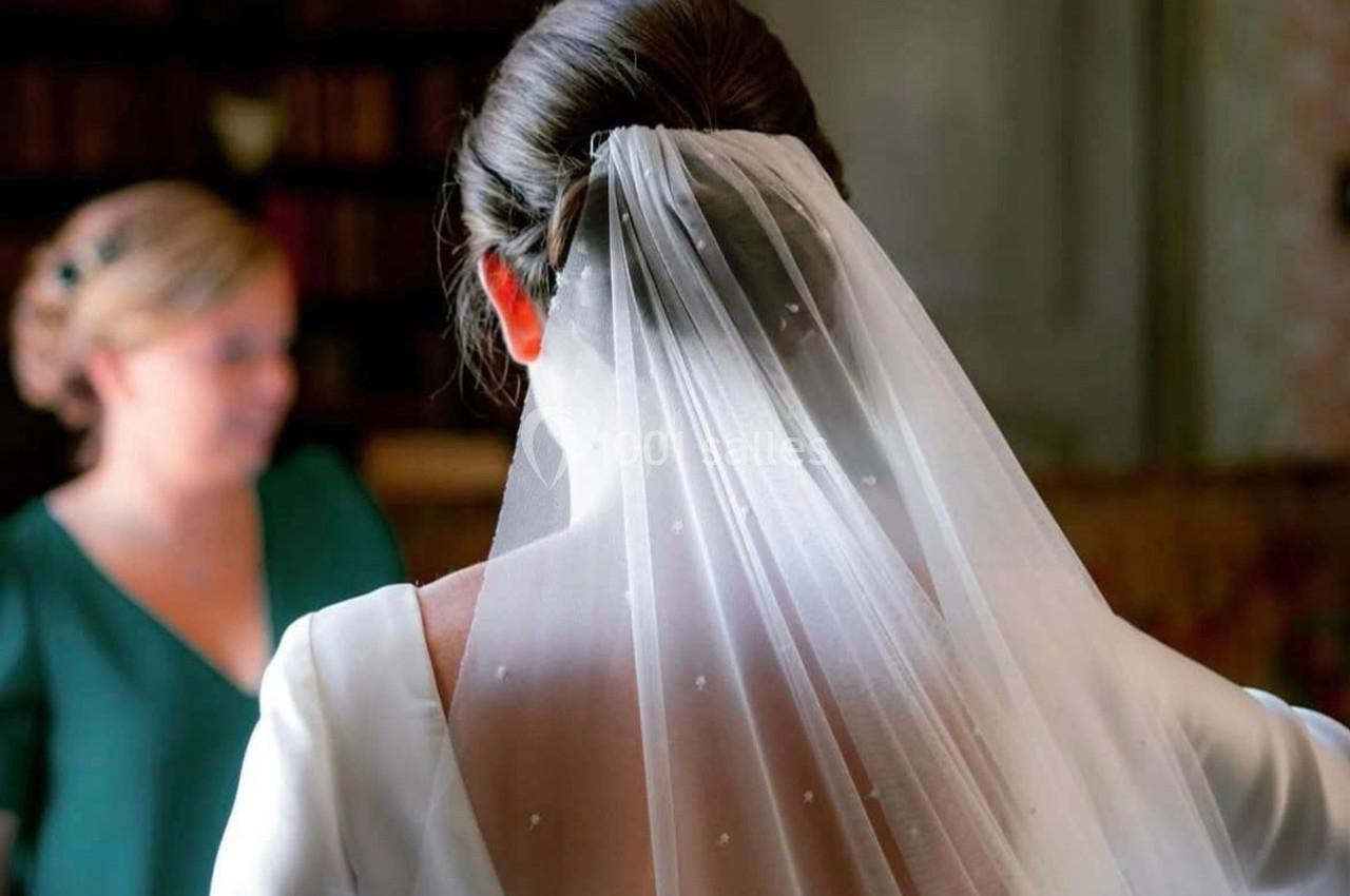 Une femme en robe de mariée vue de dos, portant un voile blanc, avec une autre personne floue en arrière-plan.