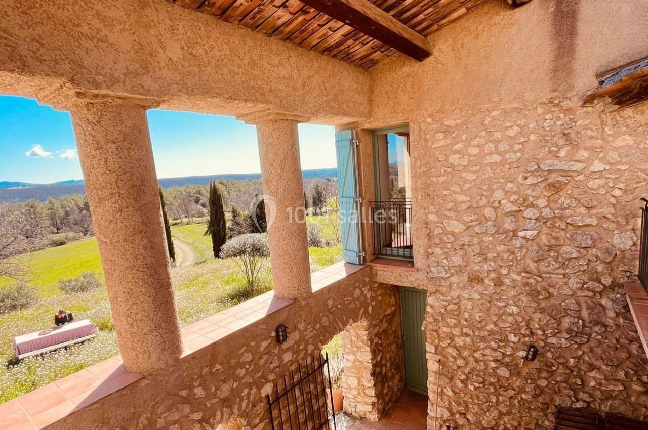 Vue d'une terrasse en pierre avec colonnes, donnant sur un paysage champêtre et vallonné sous un ciel dégagé.