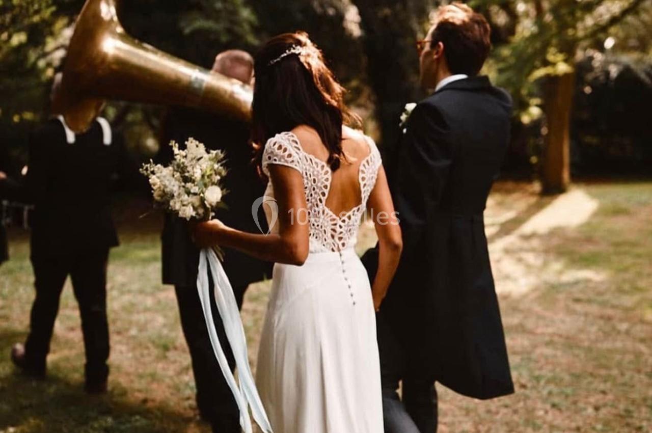 Une mariée en robe blanche et un homme en costume marchent dans un jardin, suivis par un musicien avec un tuba.