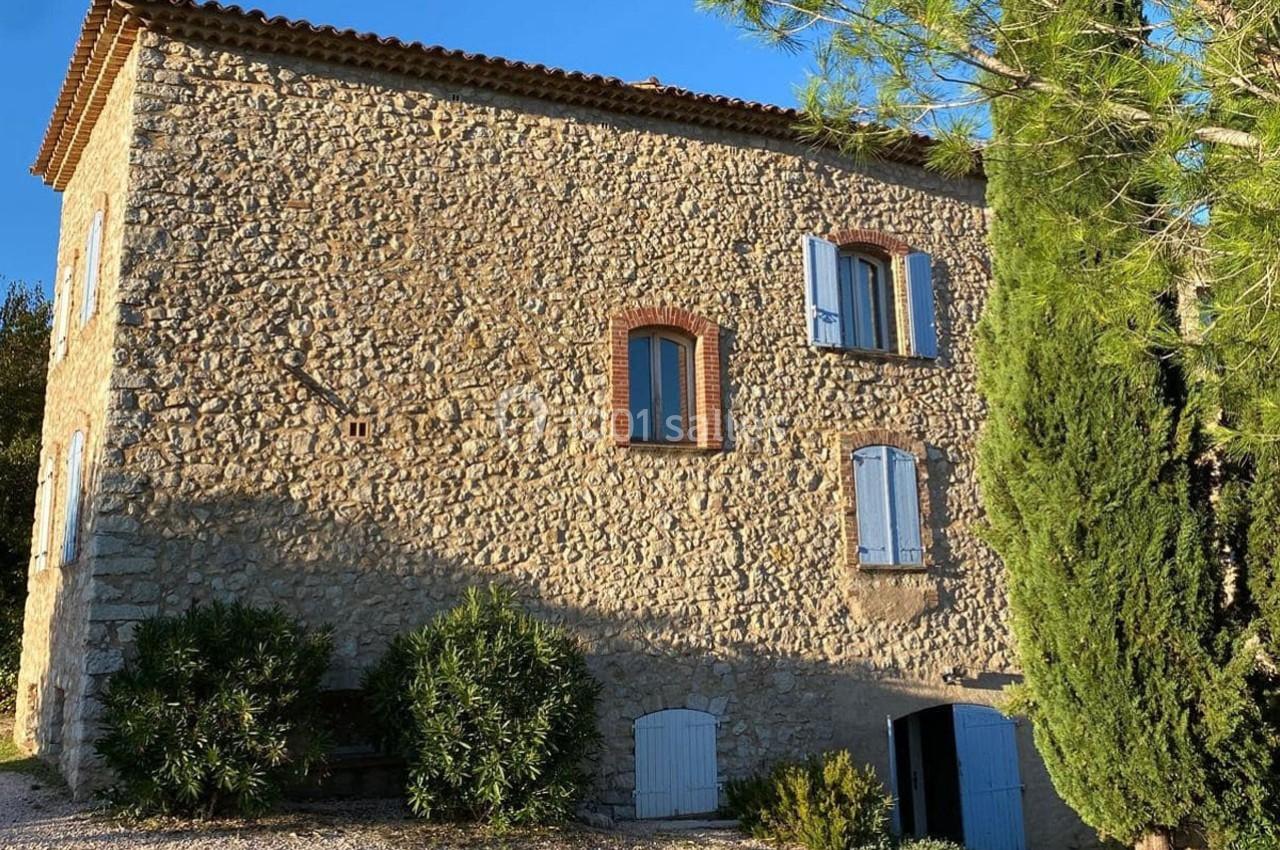 Façade en pierre d'une maison traditionnelle avec volets bleus, entourée de végétation et éclairée par le soleil.