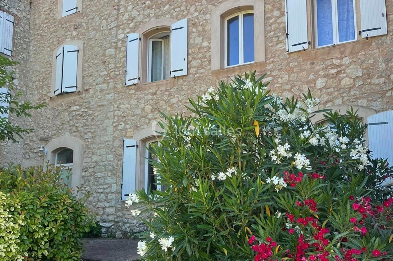 Façade en pierre d'une maison avec volets blancs, entourée de buissons fleuris rouges et blancs.