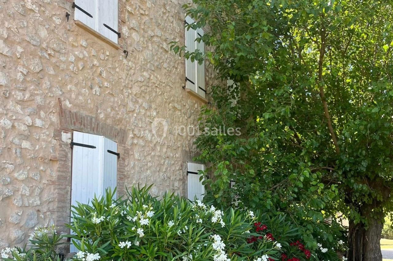 Façade en pierre d'une maison avec volets blancs, entourée de lauriers fleuris et d'un arbre verdoyant.