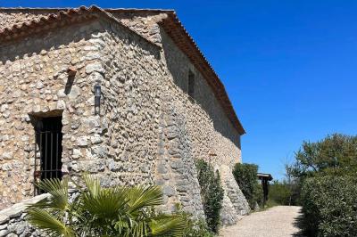 Bâtisse en pierre avec toit en tuiles, entourée de végétation et d'un chemin sous un ciel bleu clair.