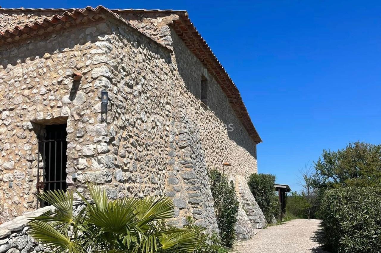 Bâtisse en pierre avec toit en tuiles, entourée de végétation et d'un chemin sous un ciel bleu clair.