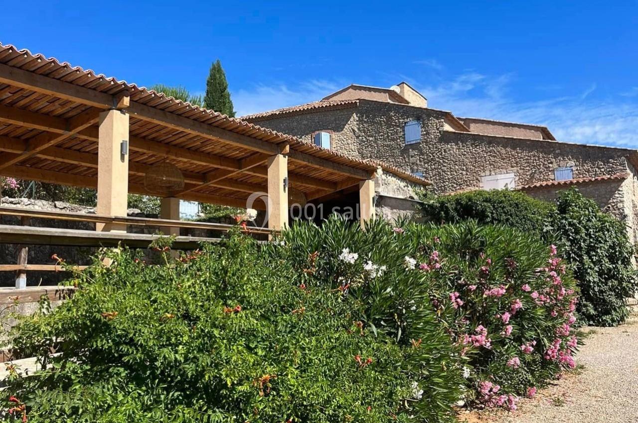 Bâtiment en pierre avec toit en tuiles, pergola en bois et jardin fleuri sous un ciel bleu.