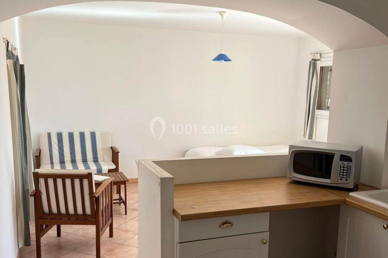 Vue d'un petit studio avec coin cuisine, canapé, table basse, lit et lumière naturelle entrant par une fenêtre.