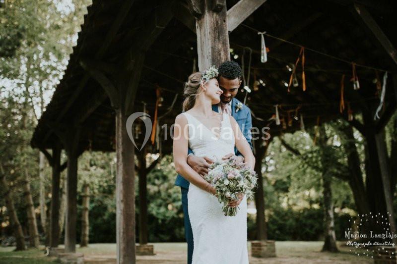 Un couple en tenue de mariage s'enlace sous une structure en bois dans un cadre extérieur verdoyant.