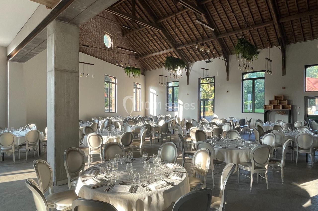 Salle de réception lumineuse avec tables rondes dressées, chaises élégantes et plafond en bois apparent.