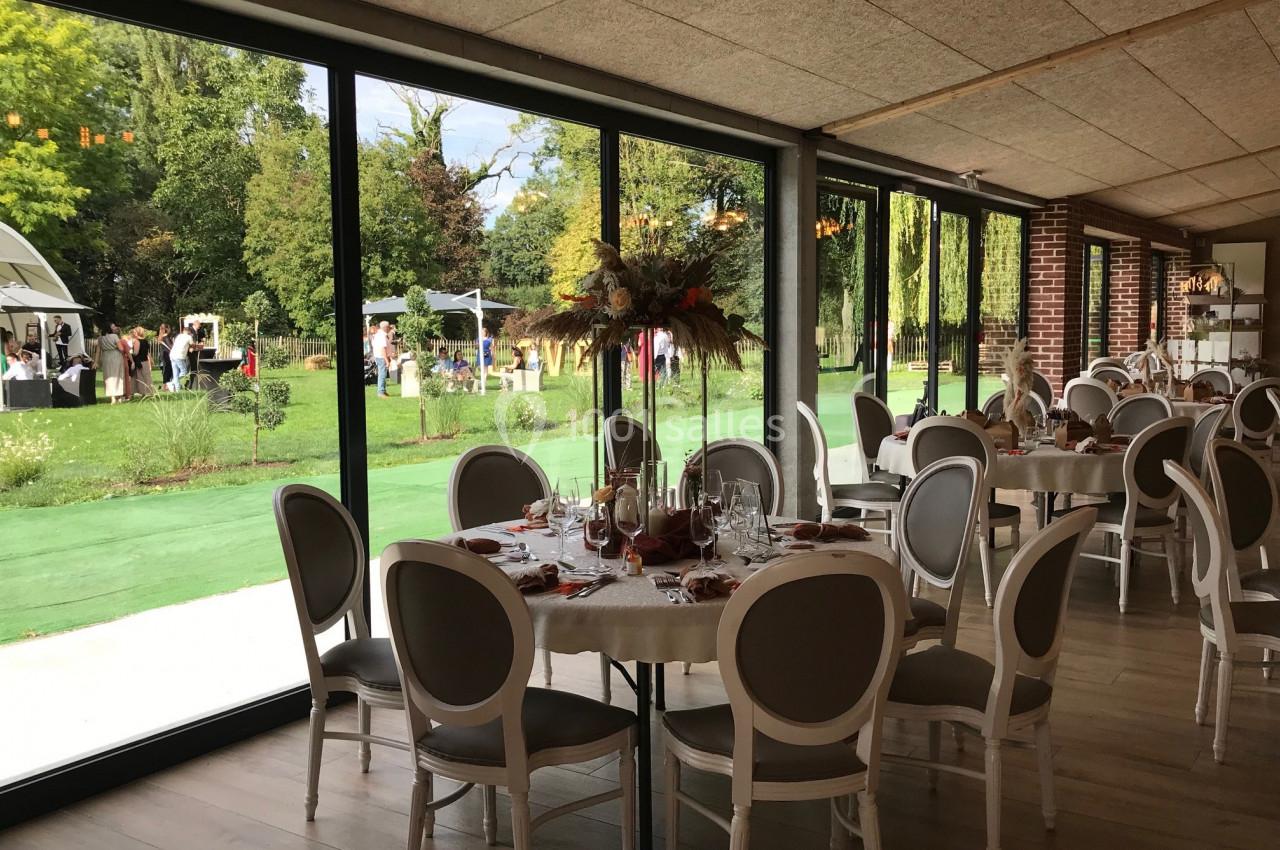 Salle de réception lumineuse avec tables dressées, vue sur un jardin verdoyant et une fête en extérieur.