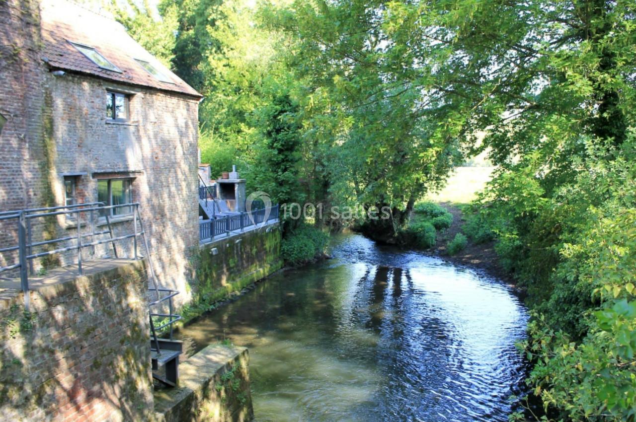 Vue d'un moulin en briques au bord d'une rivière entourée de végétation verdoyante.