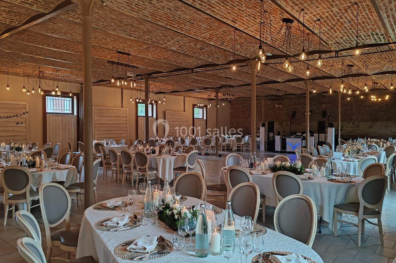 Salle de réception décorée avec des tables rondes élégantes, nappes blanches, chandeliers et éclairage suspendu.