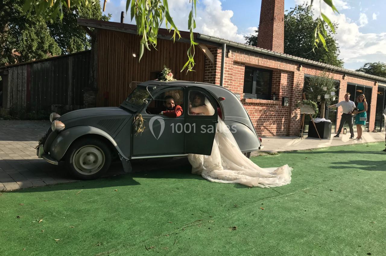 Une mariée sort d'une voiture ancienne grise devant un bâtiment en briques, sur une allée en gazon artificiel.