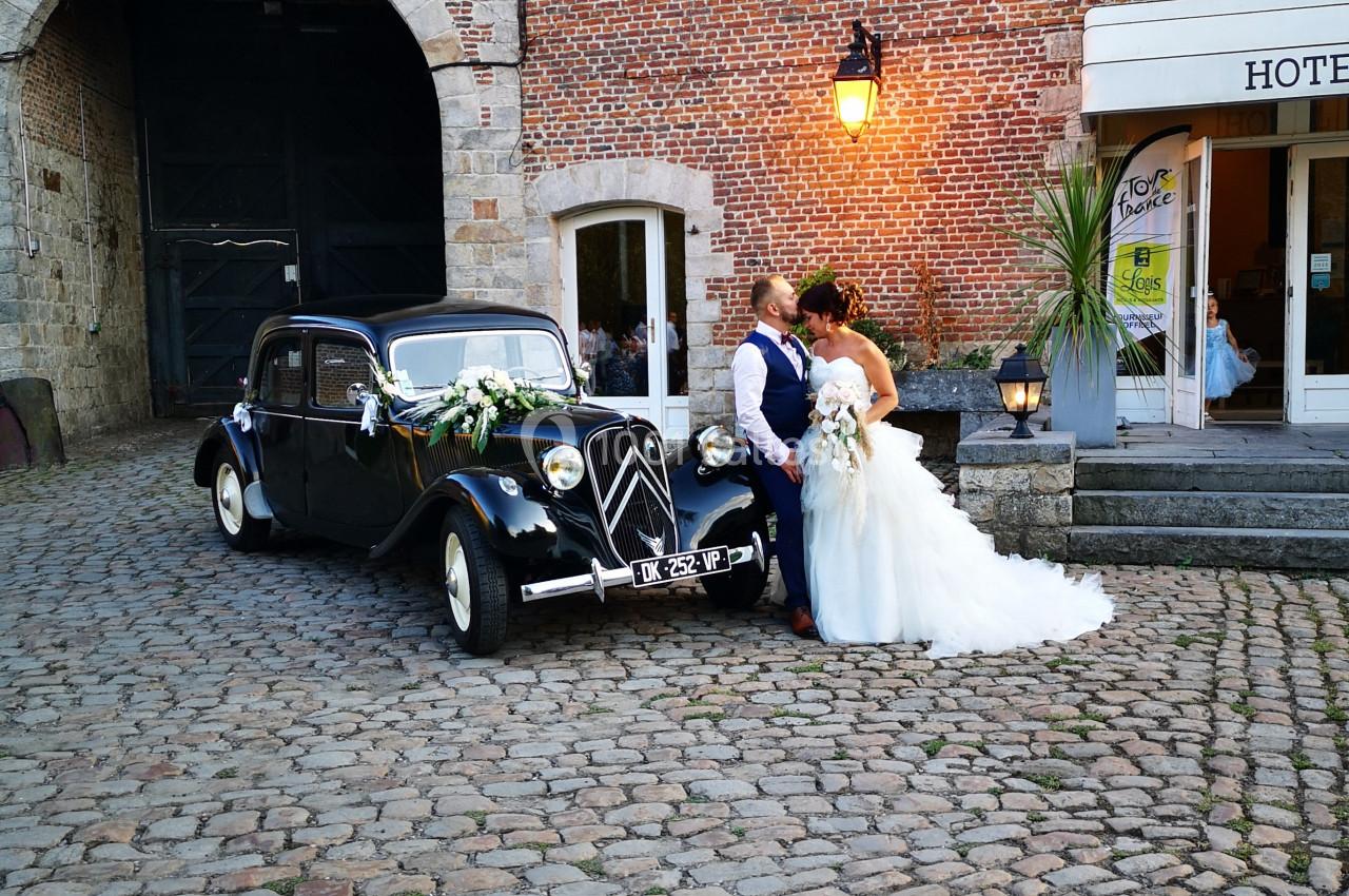 Un couple de mariés pose devant une voiture ancienne noire sur une cour pavée près d'un bâtiment en briques.