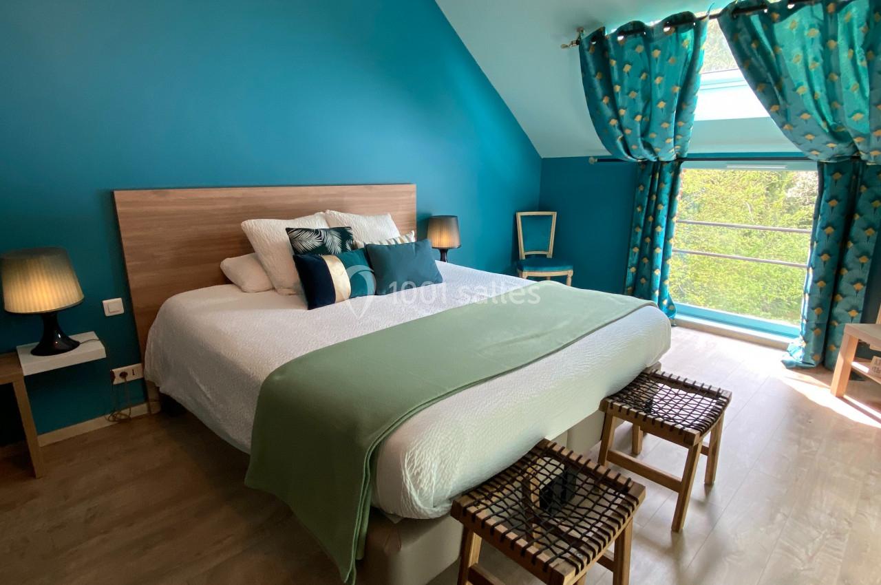 Chambre lumineuse avec lit double, murs bleus, rideaux assortis, parquet clair et vue sur un espace extérieur verdoyant.