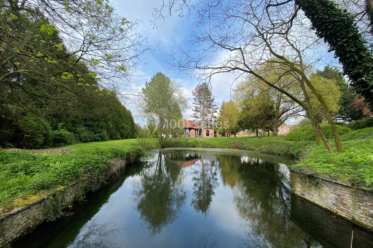 Étang entouré de verdure et d'arbres, avec un bâtiment en briques rouges visible en arrière-plan sous un ciel bleu.