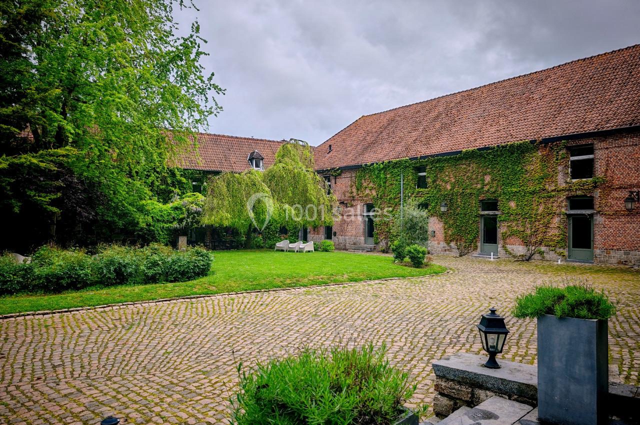Cour pavée entourée de bâtiments en briques avec toits en tuiles, végétation et bancs sous un ciel nuageux.