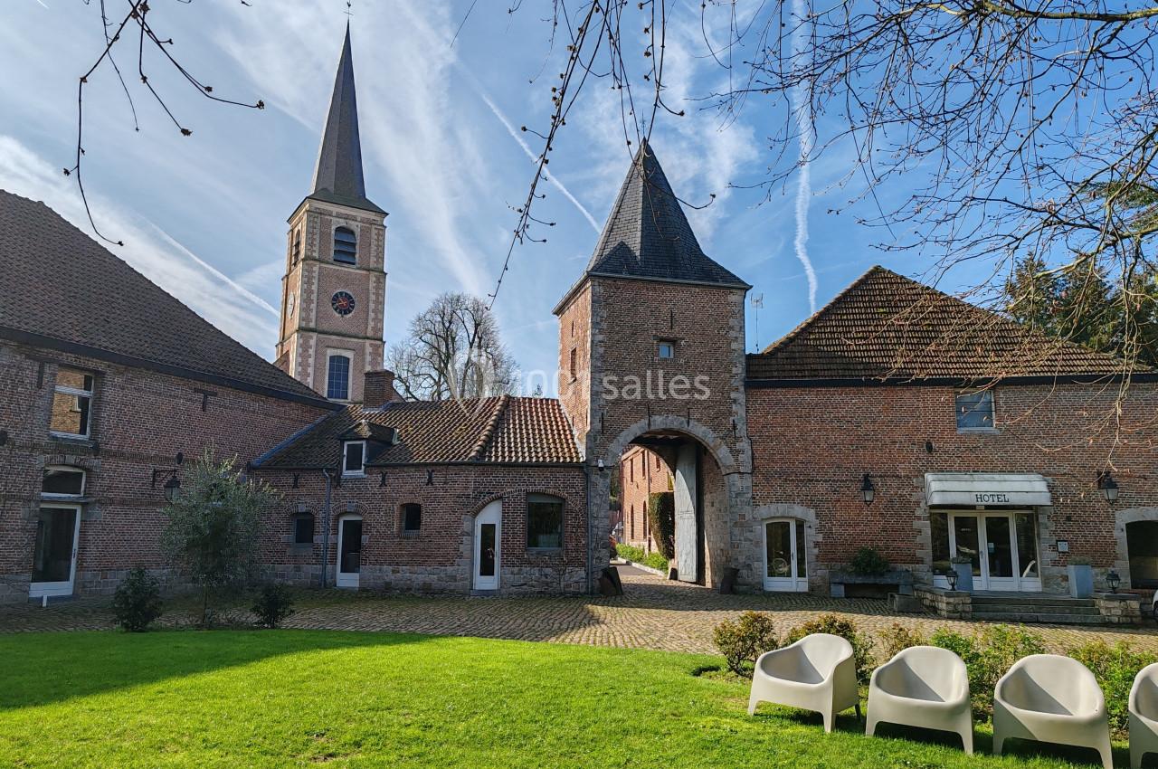 Cour intérieure d’un bâtiment en briques rouges avec une tour, une arche et des chaises blanches sur une pelouse.