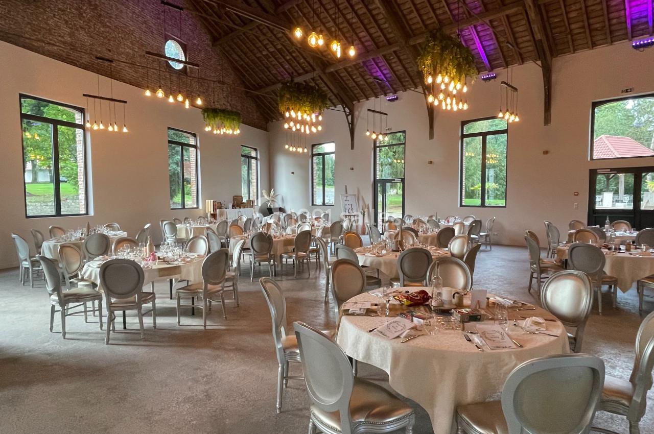Salle de réception lumineuse avec tables rondes dressées, chaises argentées et décorations suspendues sous un plafond en…