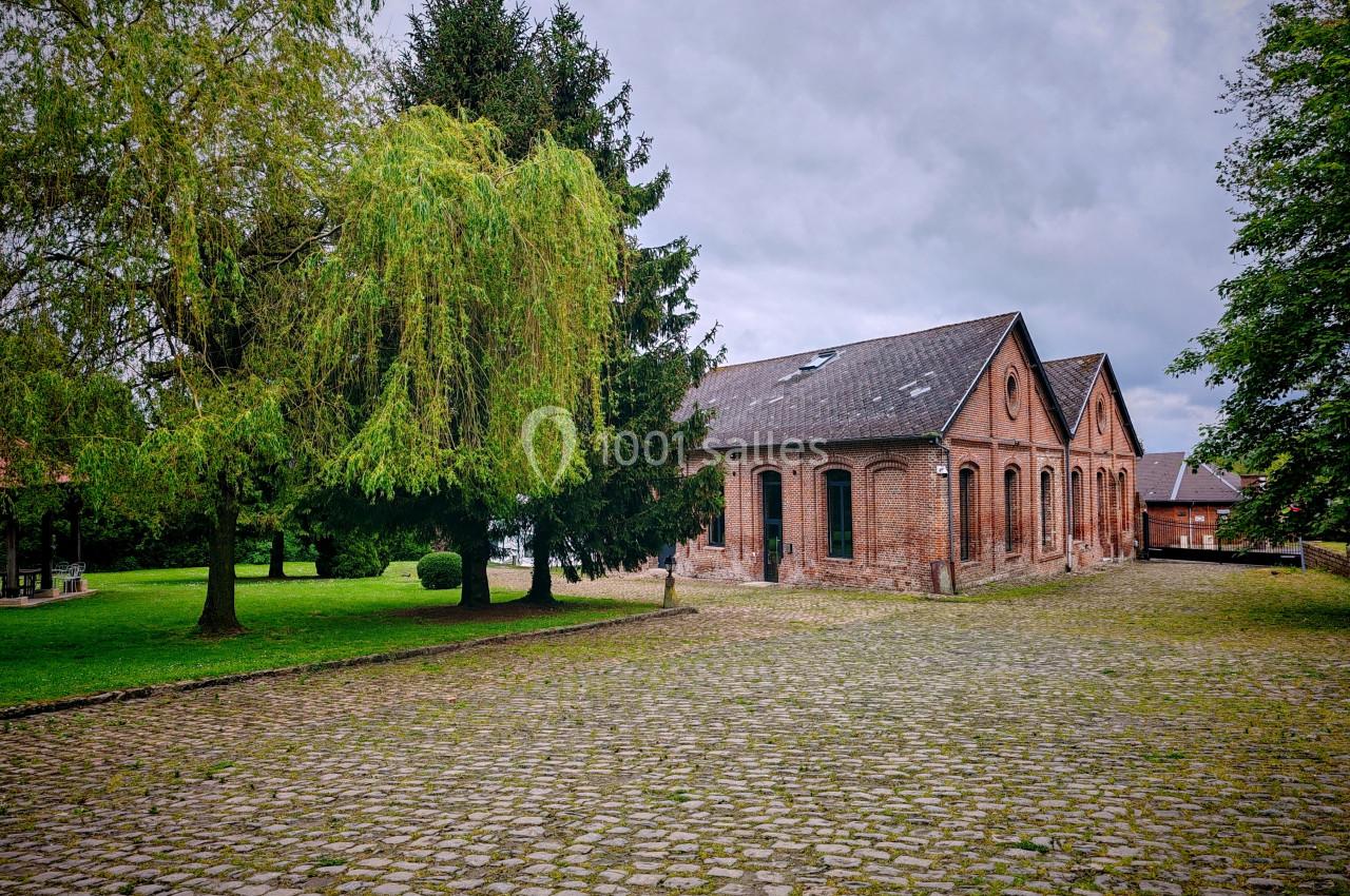 Bâtiment en briques rouges entouré d'arbres et d'une cour pavée sous un ciel nuageux.