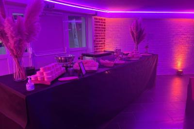 Table de buffet décorée de pampas, éclairée par des lumières violettes, avec plats et condiments disposés.