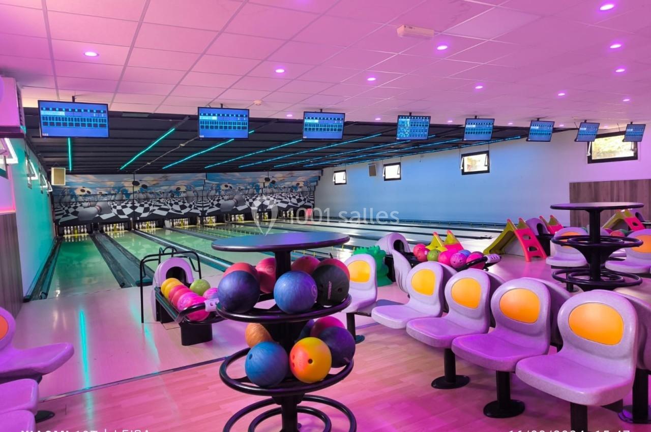 Salle de bowling avec plusieurs pistes, boules colorées sur des supports et sièges alignés sous un éclairage violet.
