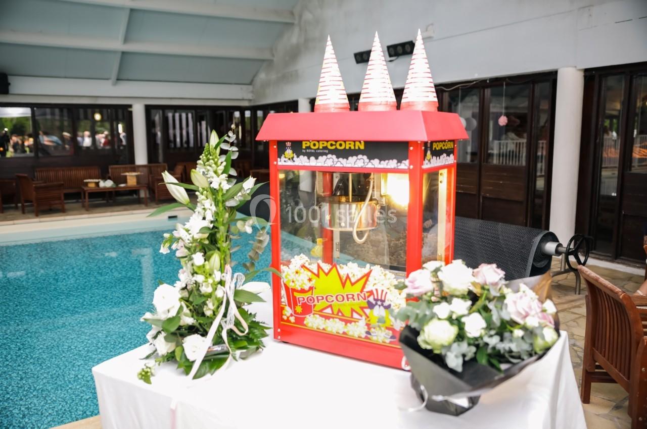 Machine à pop-corn rouge décorée, entourée de fleurs blanches et roses, placée près d'une piscine intérieure.