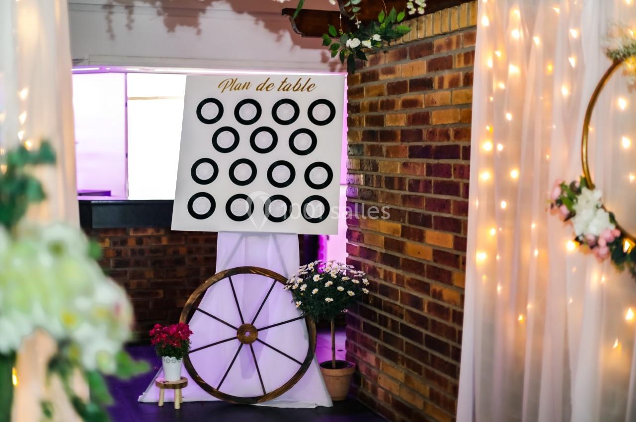Panneau de plan de table avec cercles noirs, décoré de fleurs, posé sur une roue en bois dans un espace lumineux.