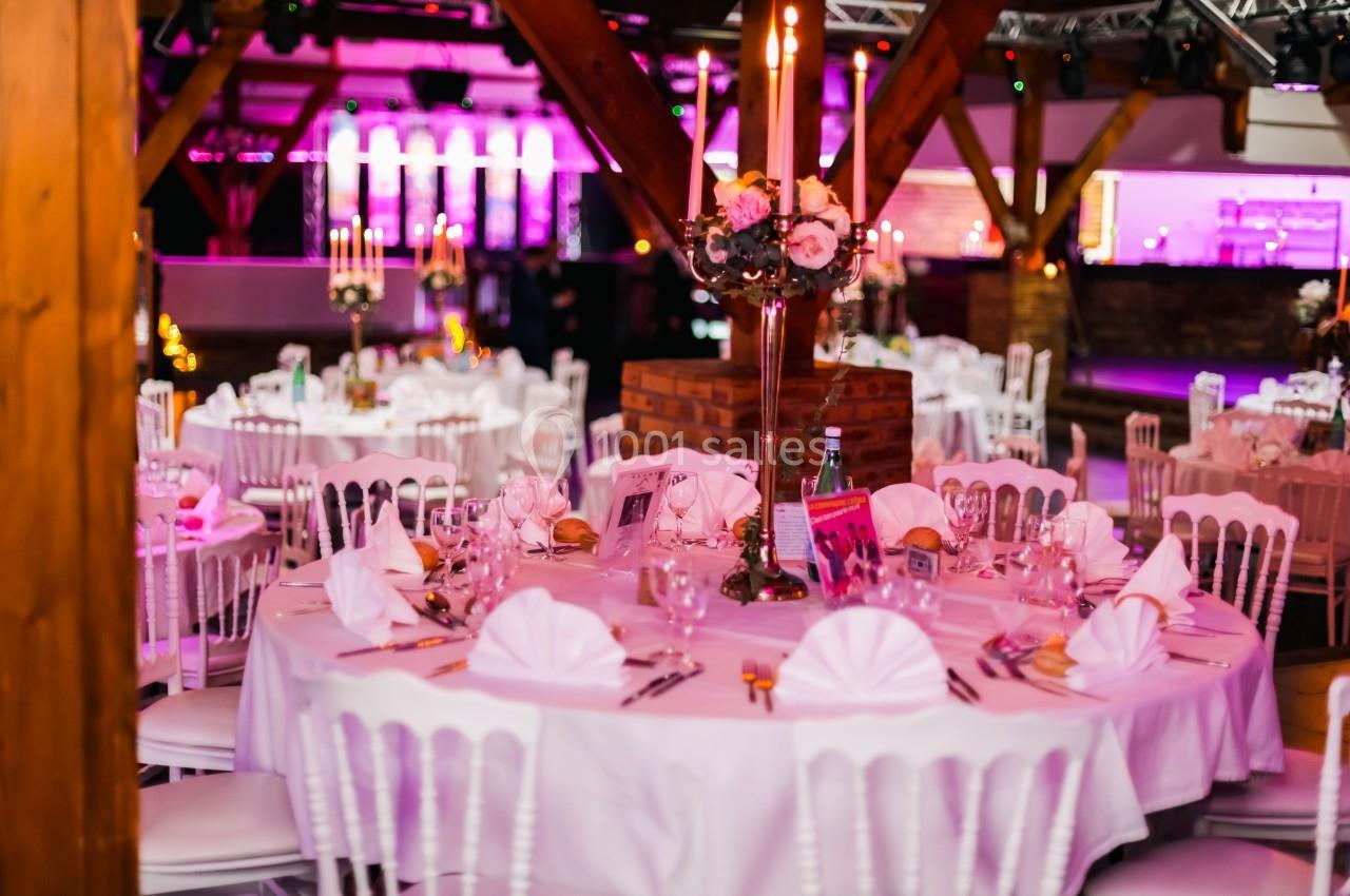 Salle de réception décorée avec des tables rondes dressées, nappes blanches, chandeliers et éclairage rose.
