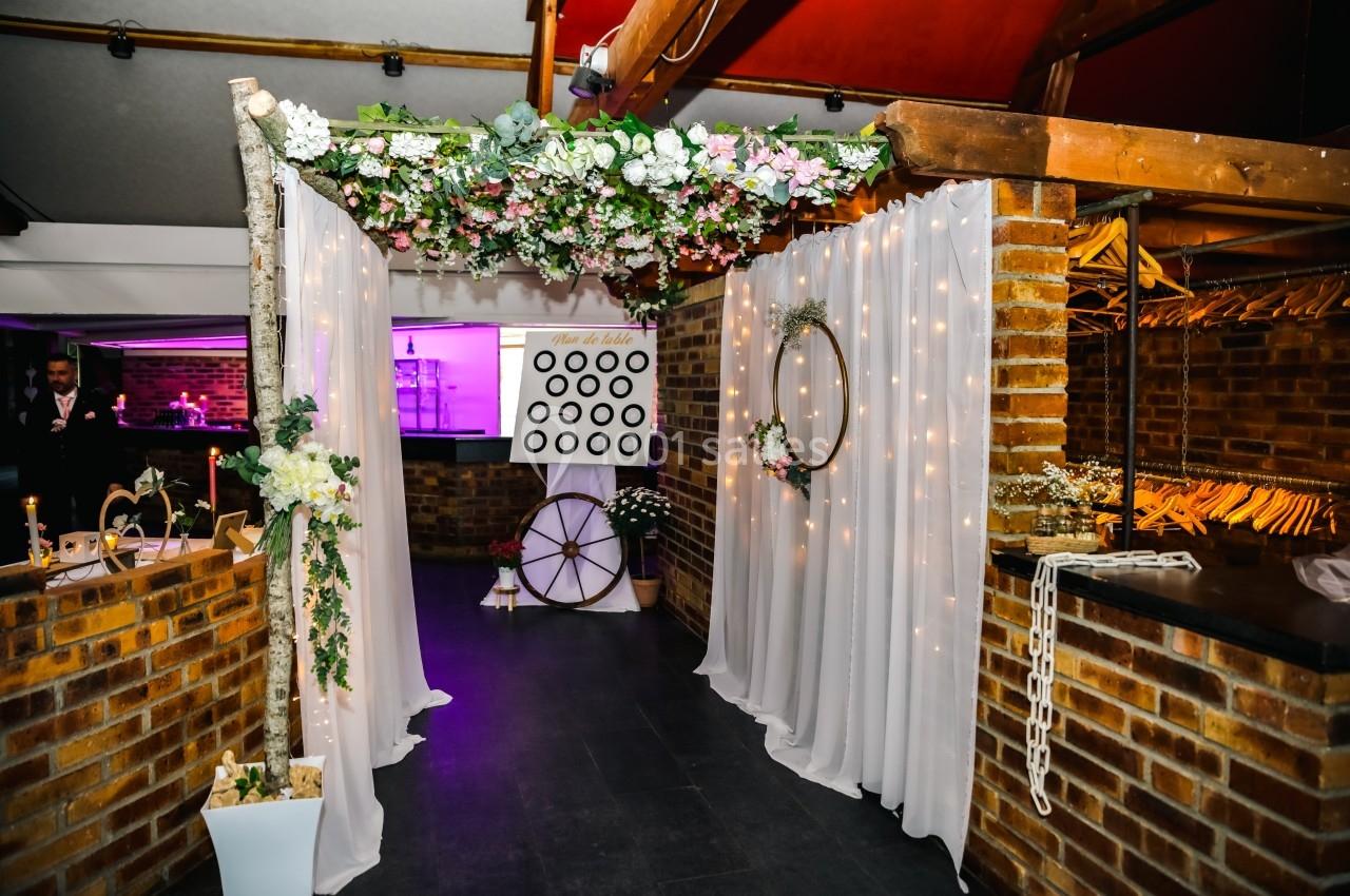 Décoration de mariage avec arche florale, rideaux blancs, guirlandes lumineuses et mur en briques dans une salle.