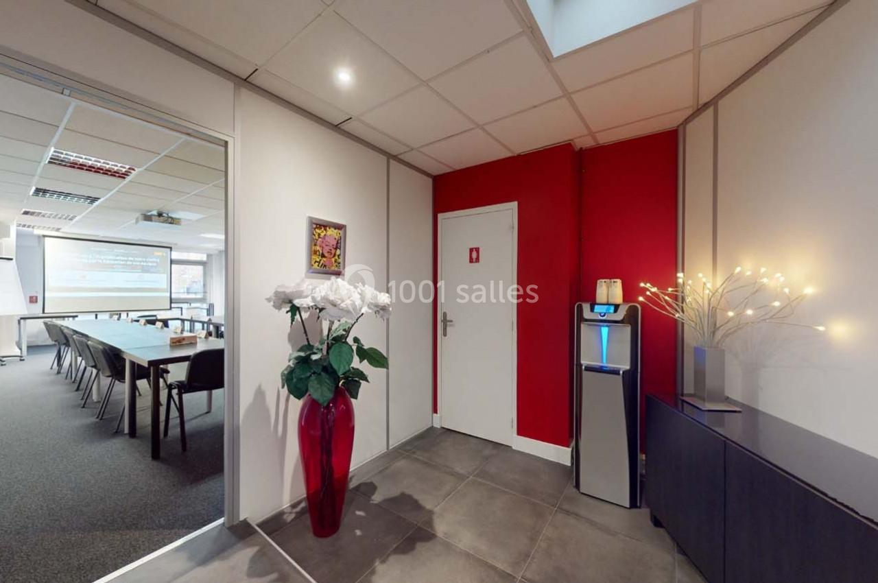 Vue d'un couloir avec une porte rouge, un distributeur d'eau, un vase de fleurs et une salle de réunion en arrière-plan.