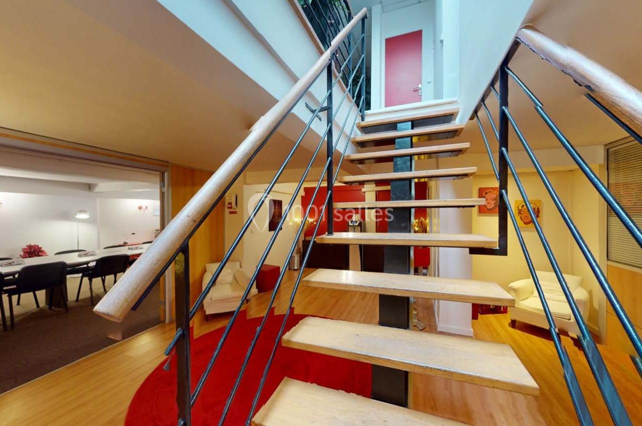 Escalier en bois avec garde-corps en métal menant à un étage, entouré d'un intérieur lumineux avec salon et salle à manger.