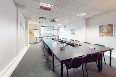 Salle de réunion avec une longue table rectangulaire, chaises noires, projecteur et murs blancs et rouges.
