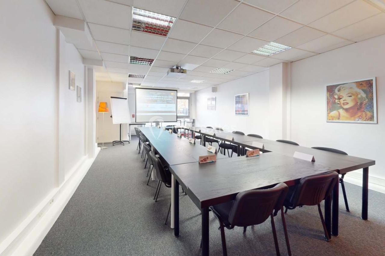 Salle de réunion lumineuse avec une grande table rectangulaire, des chaises, un projecteur et des affiches murales.