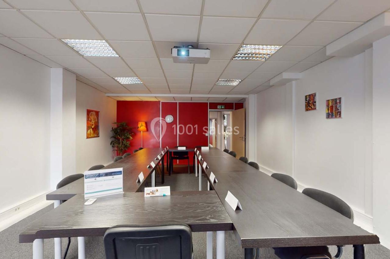 Salle de réunion lumineuse avec tables noires disposées en U, chaises noires et un projecteur fixé au plafond.