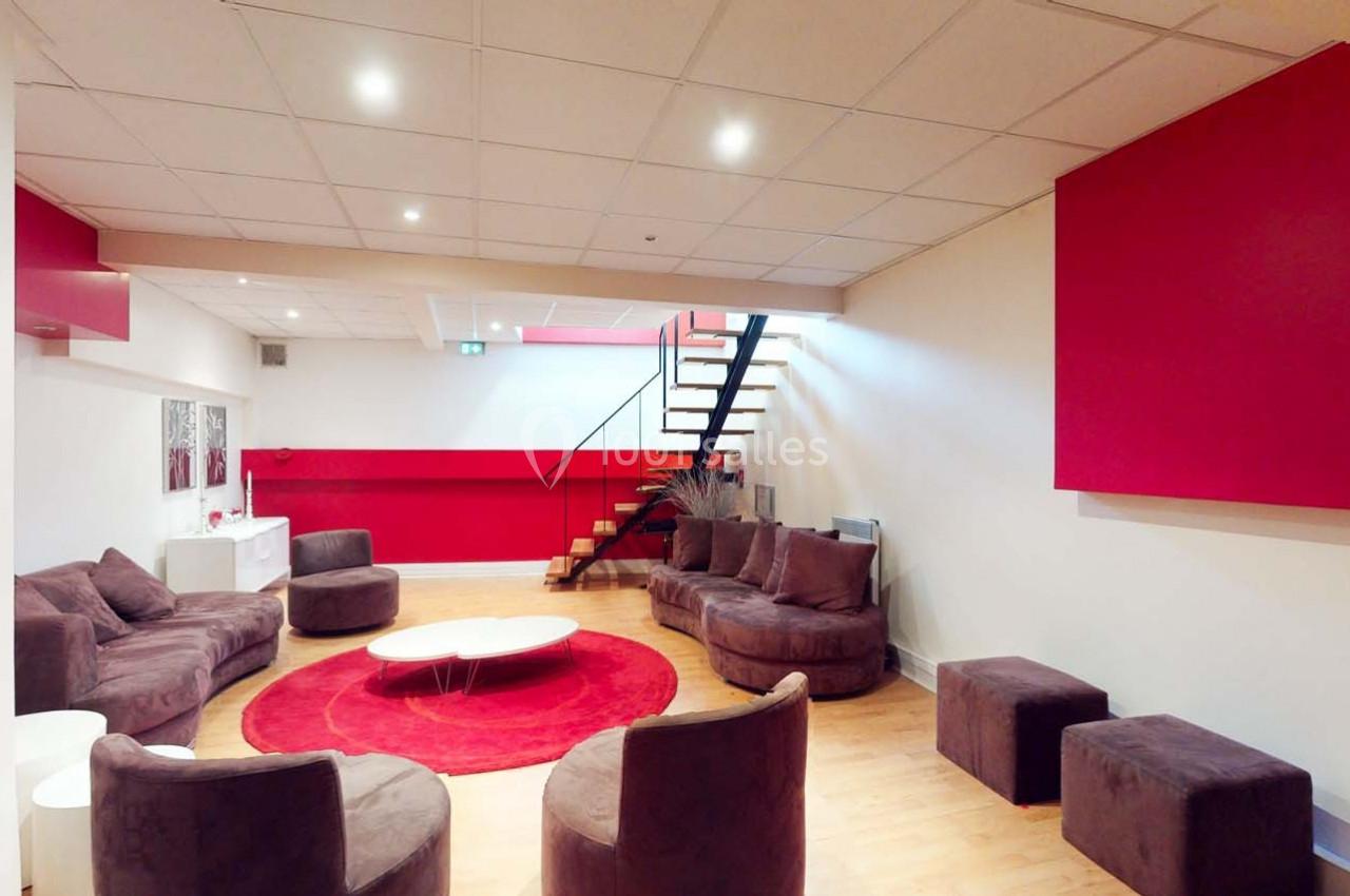 Espace salon moderne avec canapés marron, tapis rouge, escalier en métal et murs partiellement rouges.