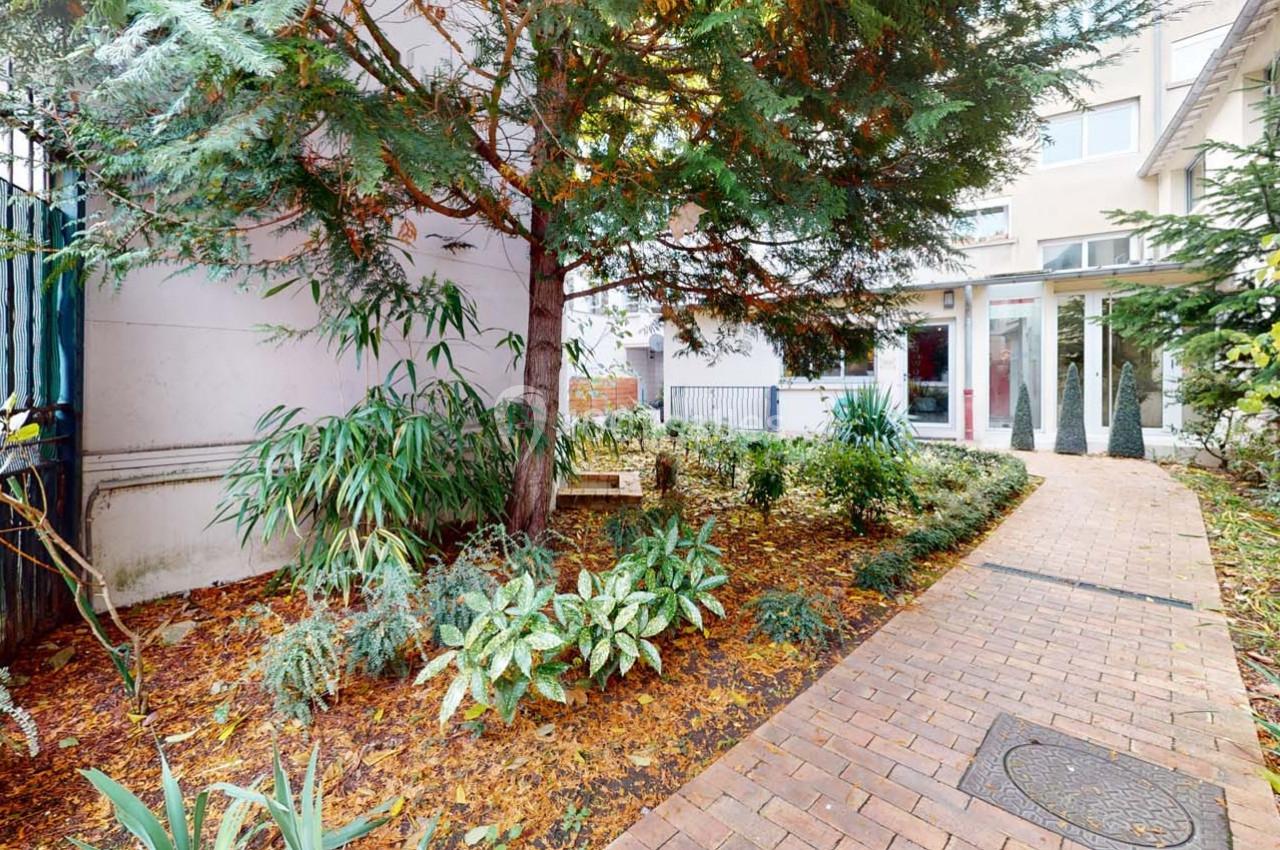 Chemin pavé bordé de plantes et d'arbustes, menant à l'entrée d'un bâtiment moderne.