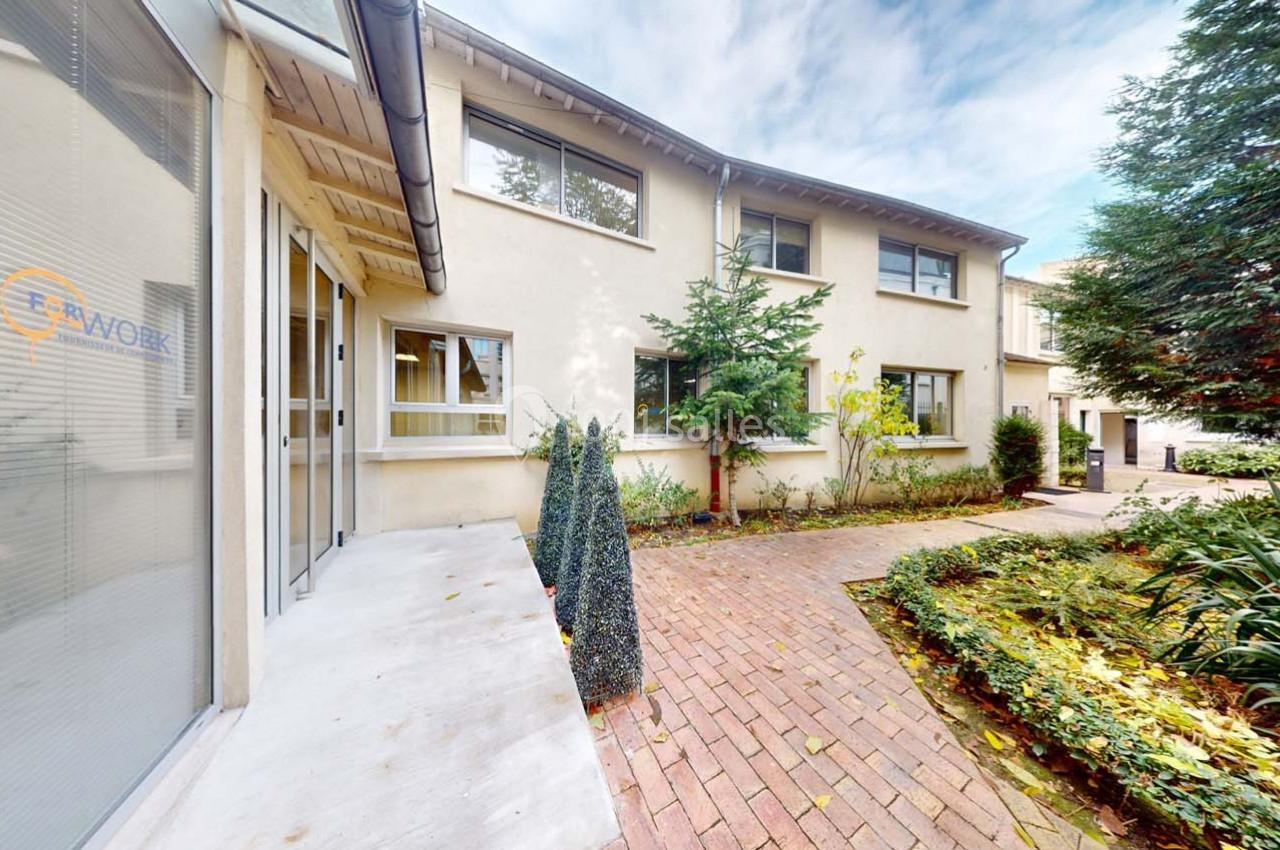 Entrée d'un bâtiment moderne entourée d'un jardin avec allée pavée et végétation.