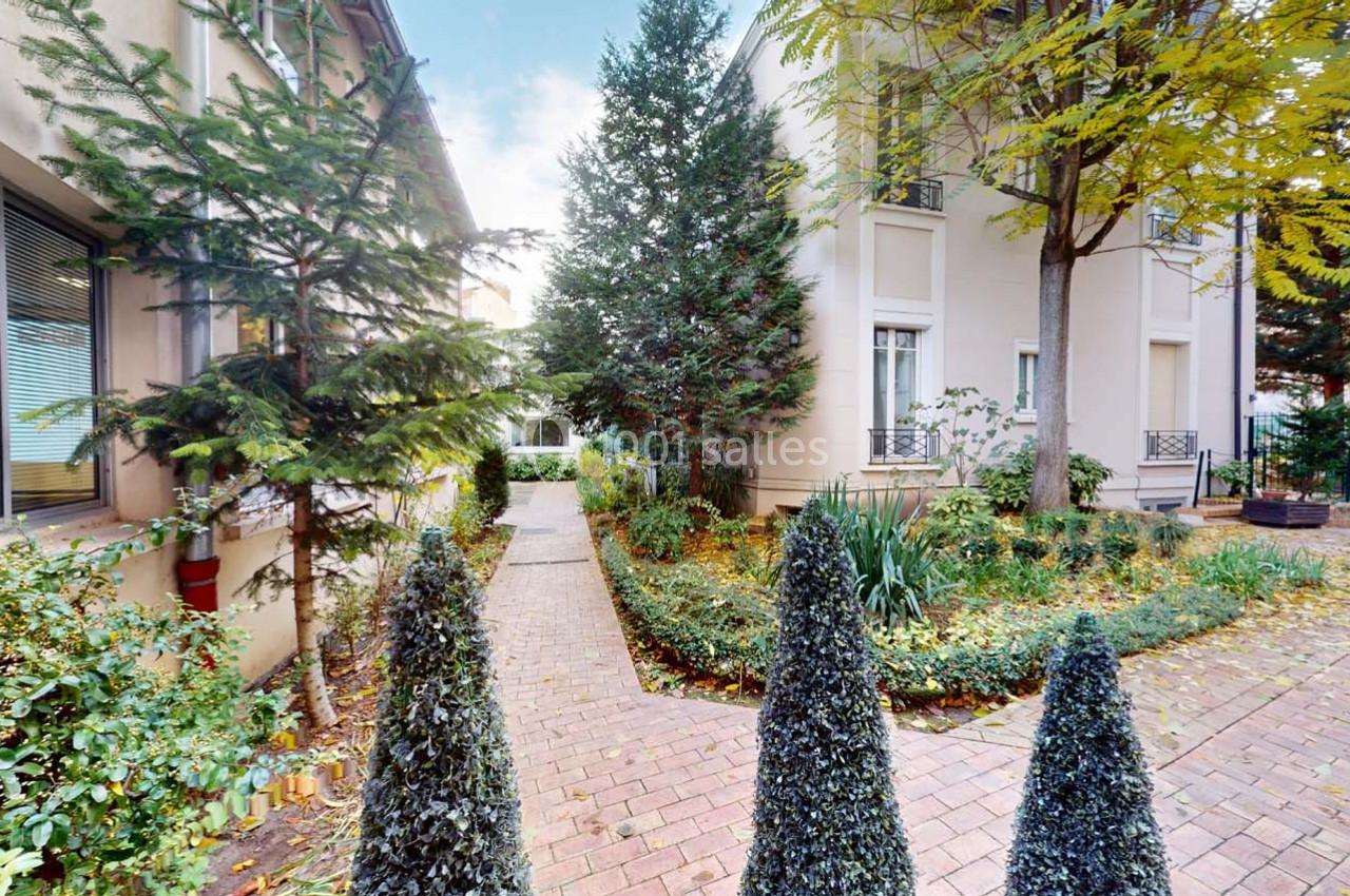 Chemin pavé bordé de végétation menant à des bâtiments résidentiels entourés d'arbres et d'arbustes.