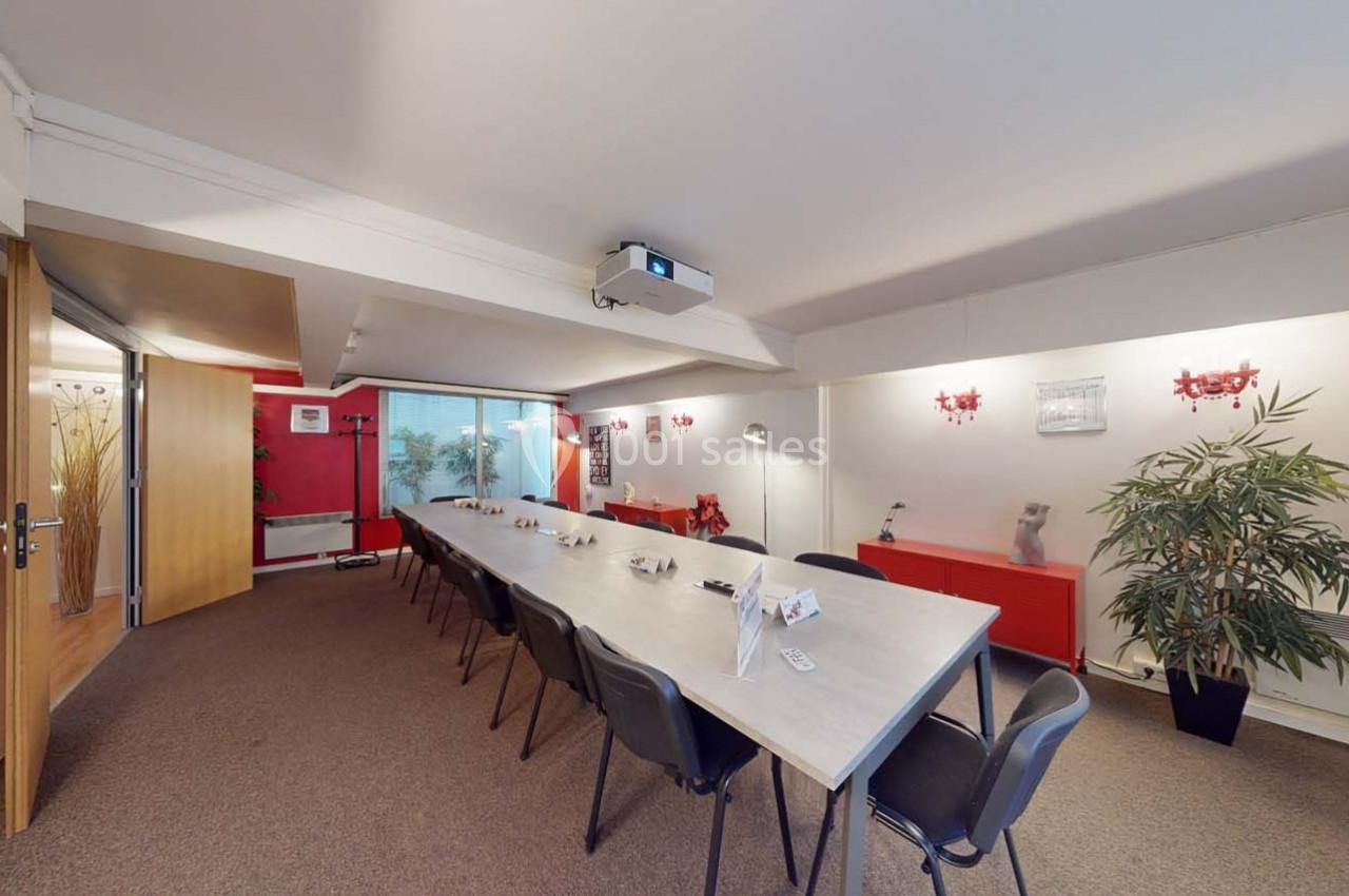 Salle de réunion lumineuse avec une grande table rectangulaire, des chaises noires et un projecteur au plafond.