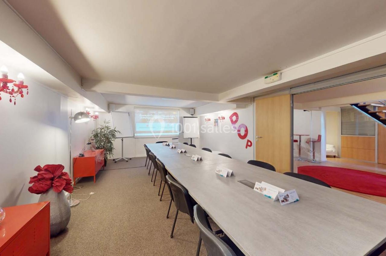 Salle de réunion lumineuse avec une grande table rectangulaire, chaises noires et écran de projection en fond.