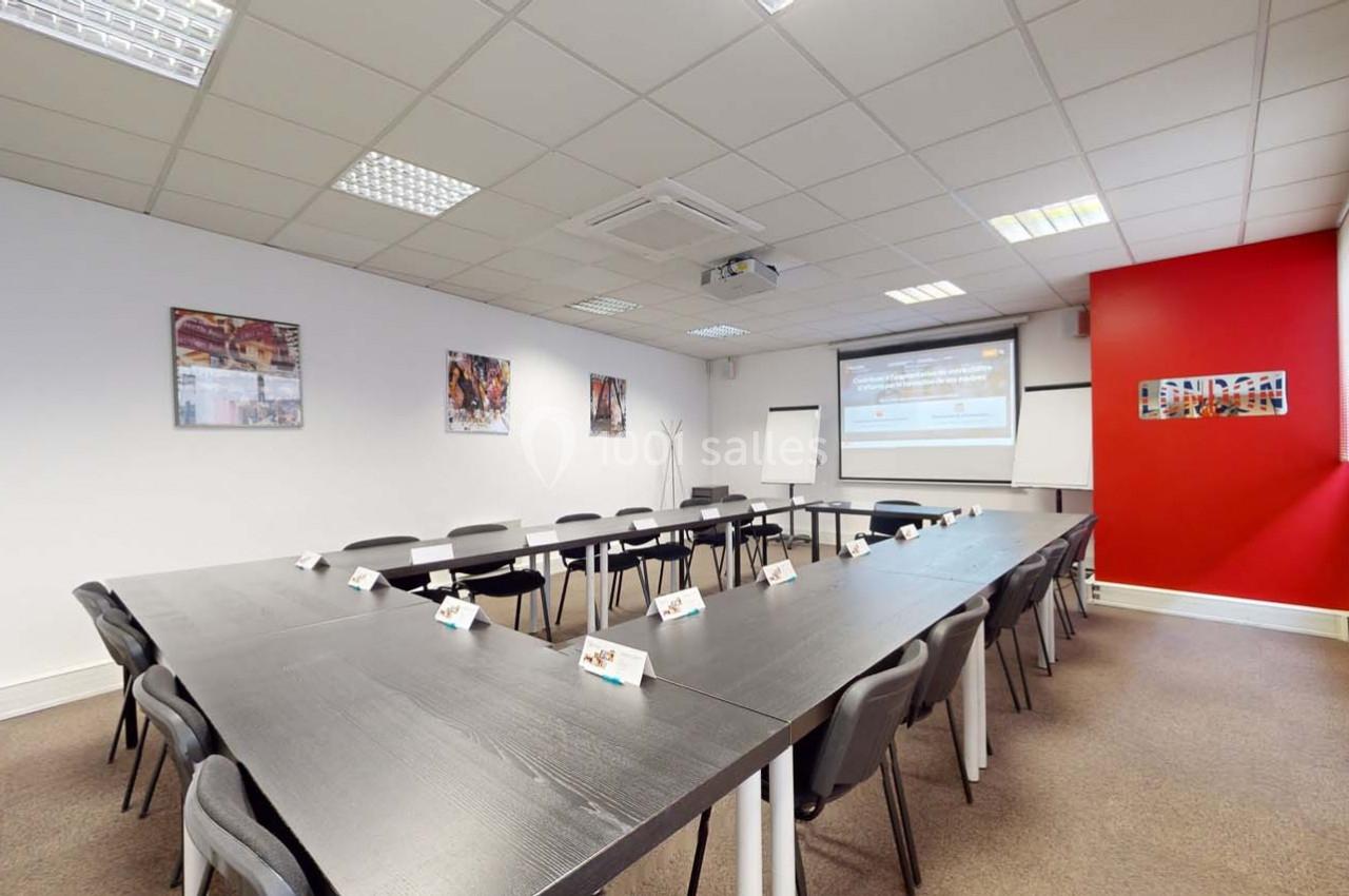 Salle de réunion lumineuse avec tables en U, chaises, écran de projection, paperboard et décorations murales.