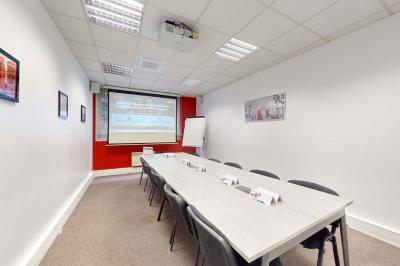 Salle de réunion avec une longue table rectangulaire, chaises noires, projecteur et murs blancs et rouges.