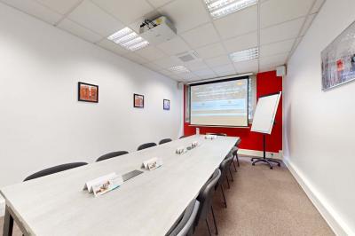 Salle de réunion avec une longue table rectangulaire, chaises noires, projecteur et murs blancs et rouges.