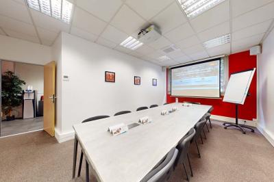 Salle de réunion avec une longue table rectangulaire, chaises noires, projecteur et murs blancs et rouges.