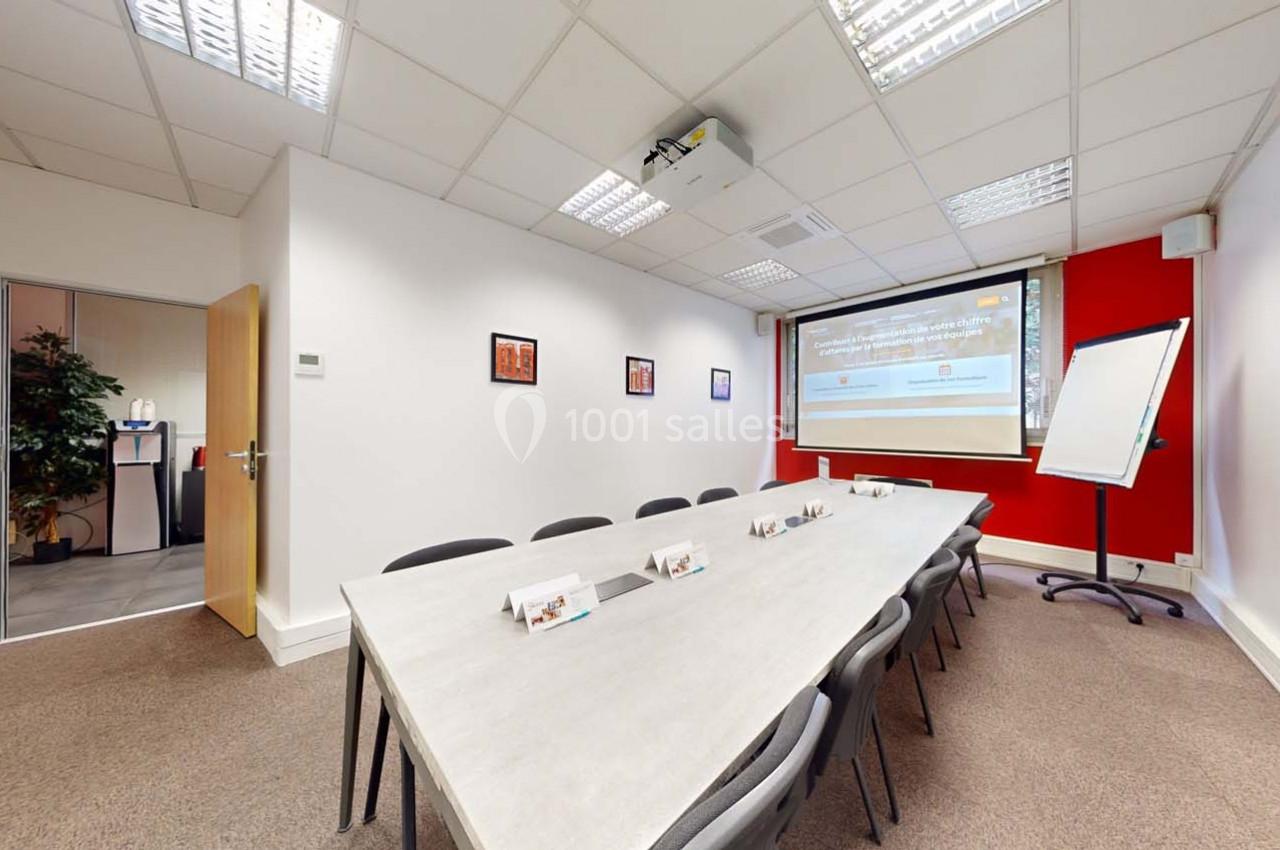 Salle de réunion avec une grande table rectangulaire, chaises, tableau blanc, projecteur et écran mural.