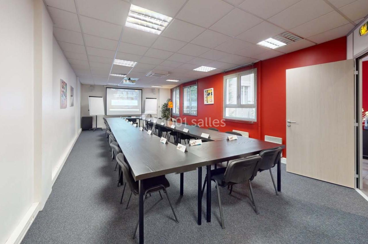 Salle de réunion avec une longue table rectangulaire, chaises noires, projecteur et murs blancs et rouges.