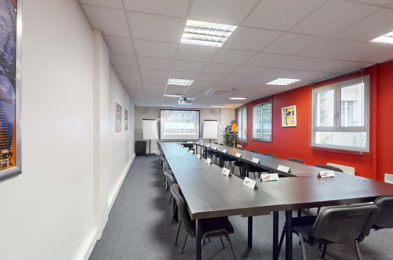 Salle de réunion lumineuse avec une longue table, des chaises, un projecteur et des murs blancs et rouges.
